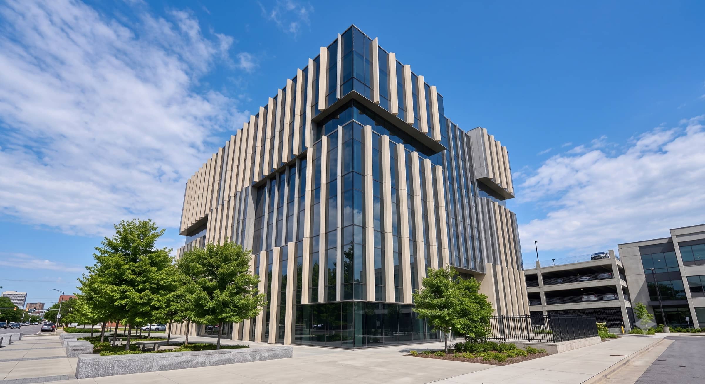 A completed multi-storey office building seen from its street corner, precast fins running the full height of two glazed elevations, plaza and young trees at its base under an open sky.